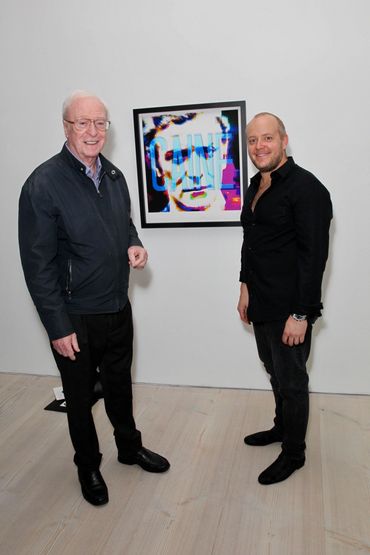 Two men standing beside a colorful framed artwork titled 'CAINE' on a white wall.