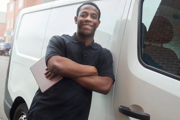 Smiling man in black shirt leaning against white van with laptop.