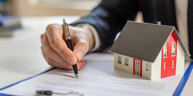 Mano con pluma firmado documentos, mostrando una casa. Hace referencia a la adquisición de inmuebles