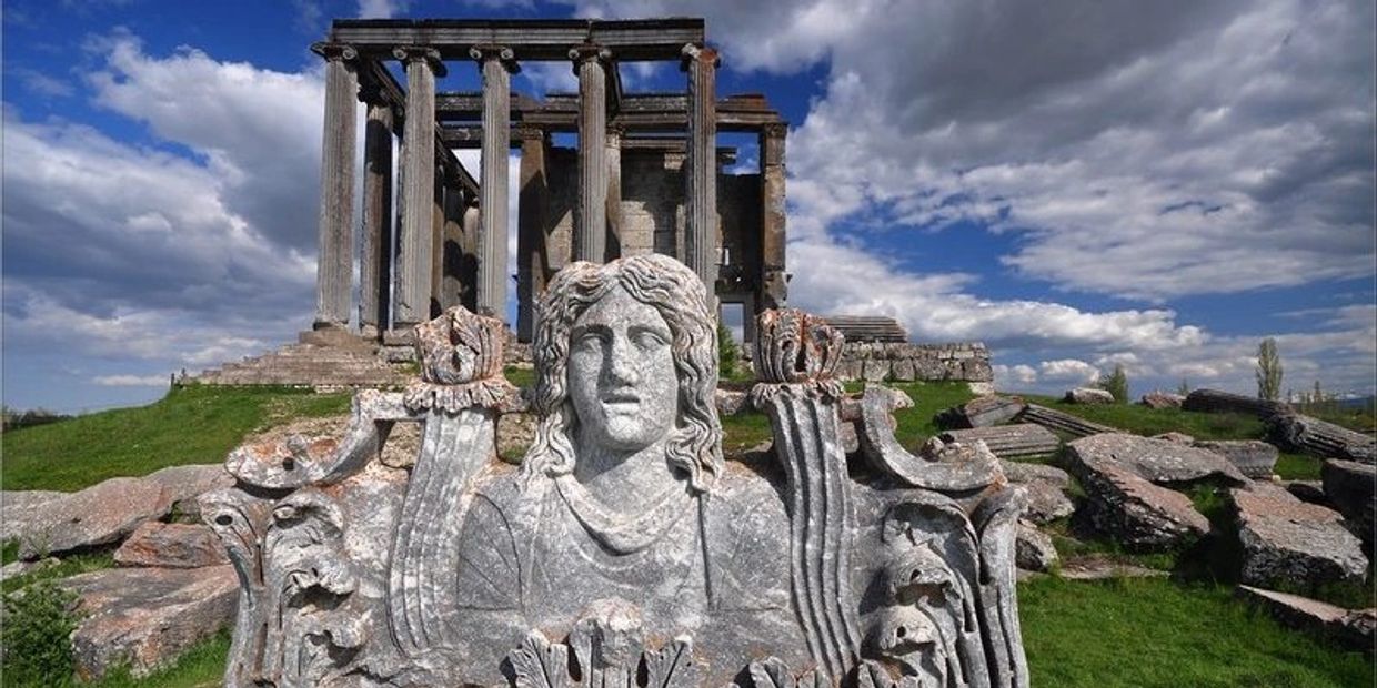 Ancient stone statue with a temple ruin under a cloudy sky.