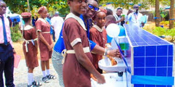 Children in school uniforms washing hands at a blue tiled handwashing station outdoors.