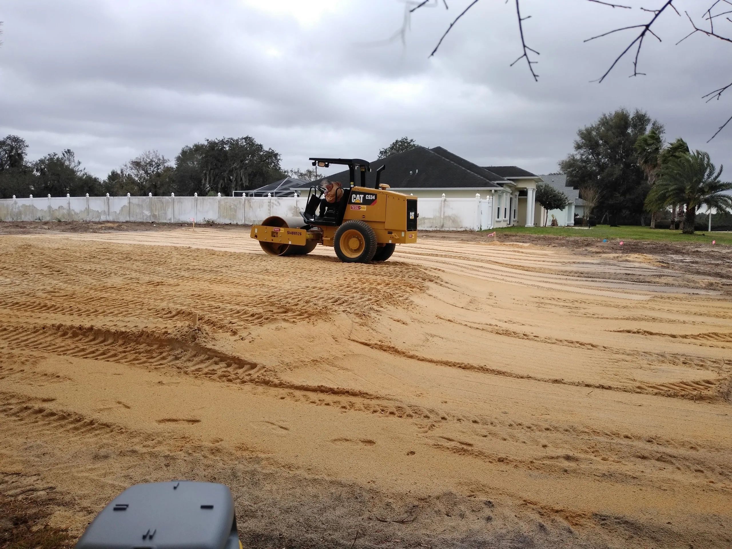 A yellow CAT CS34 roller compacts sand at a construction site near houses.