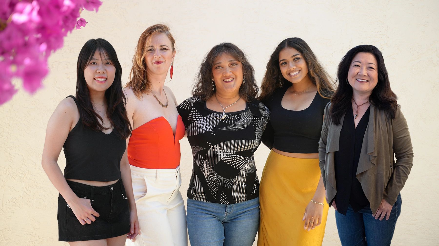 Five diverse women smiling and posing together against a light wall with pink flowers.