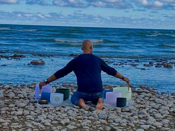 Man playing crystal singing bowls by the ocean on rocky shore.