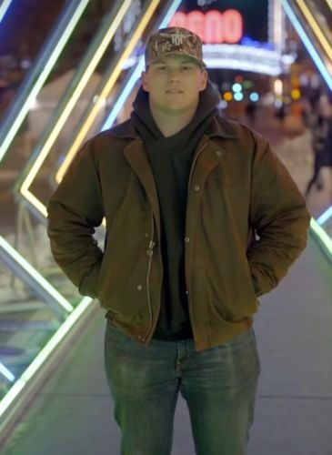 Young man in a brown jacket and camo hat standing under neon lights at night.