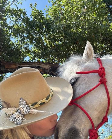 Woman wearing a decorated hat kisses a white horse with a red halter.
