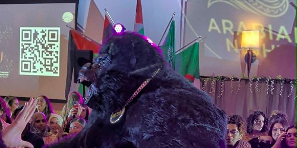 A large black bear entertaining a crowd at an indoor event with flags and a QR code in the background.
