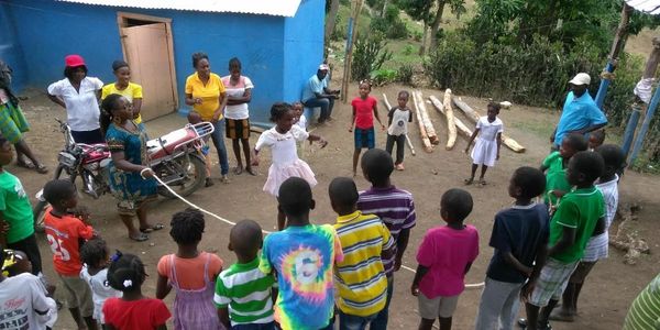 Children and adults play jump rope outside a blue building in a rural setting.