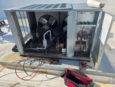 Open HVAC unit with tools and gauges on a rooftop under bright sunlight.