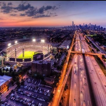 A brightly lit stadium near a busy highway at sunset with a city skyline in the background.