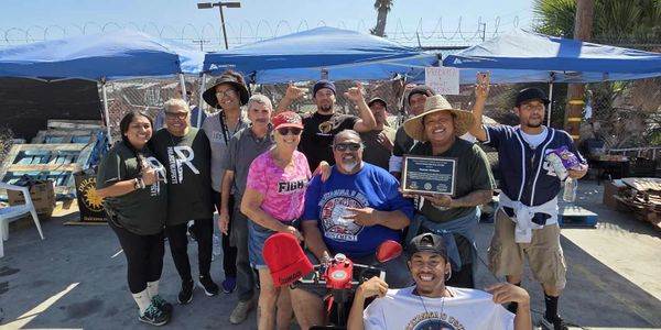 Group of people smiling outdoors under tents, celebrating with a plaque and a shirt.