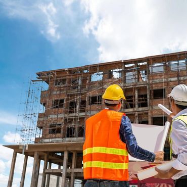 Two construction workers review blueprints at a building site under clear skies.