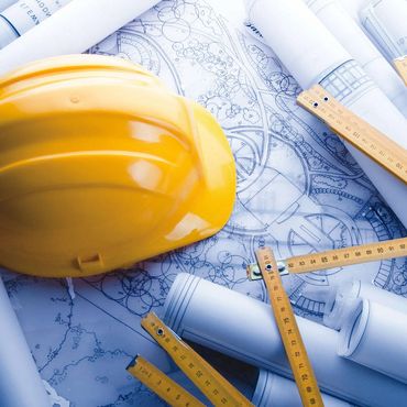 Yellow construction helmet on rolled architectural blueprints with measuring rulers.