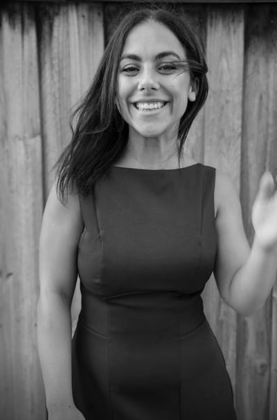 Smiling woman in a sleeveless dress stands in front of a wooden fence.