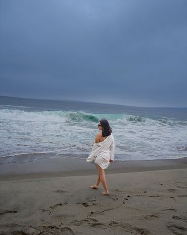 A woman in a white dress walks barefoot along a cloudy beach shore.