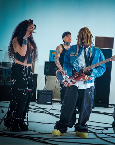 Three musicians performing with tattoos, stylish outfits, and a guitar in a studio setting.