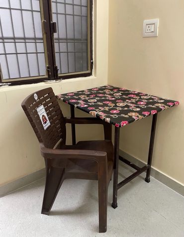 Brown plastic chair beside a floral-covered table in a corner by a window.