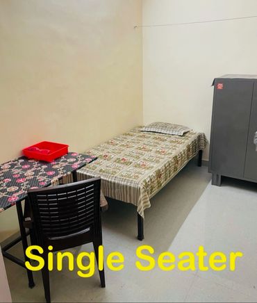 A simple single seater room with bed, chair, table, and cupboard.