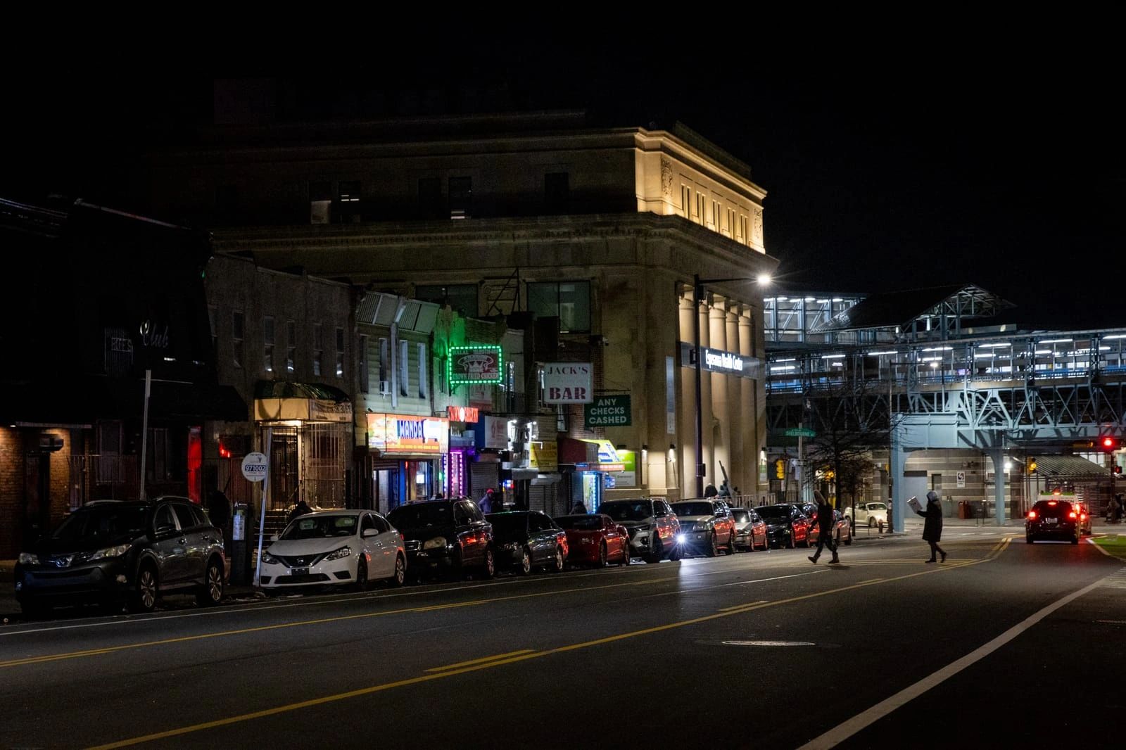 Nighttime street scene with parked cars and pedestrians near a lit building and elevated train station.