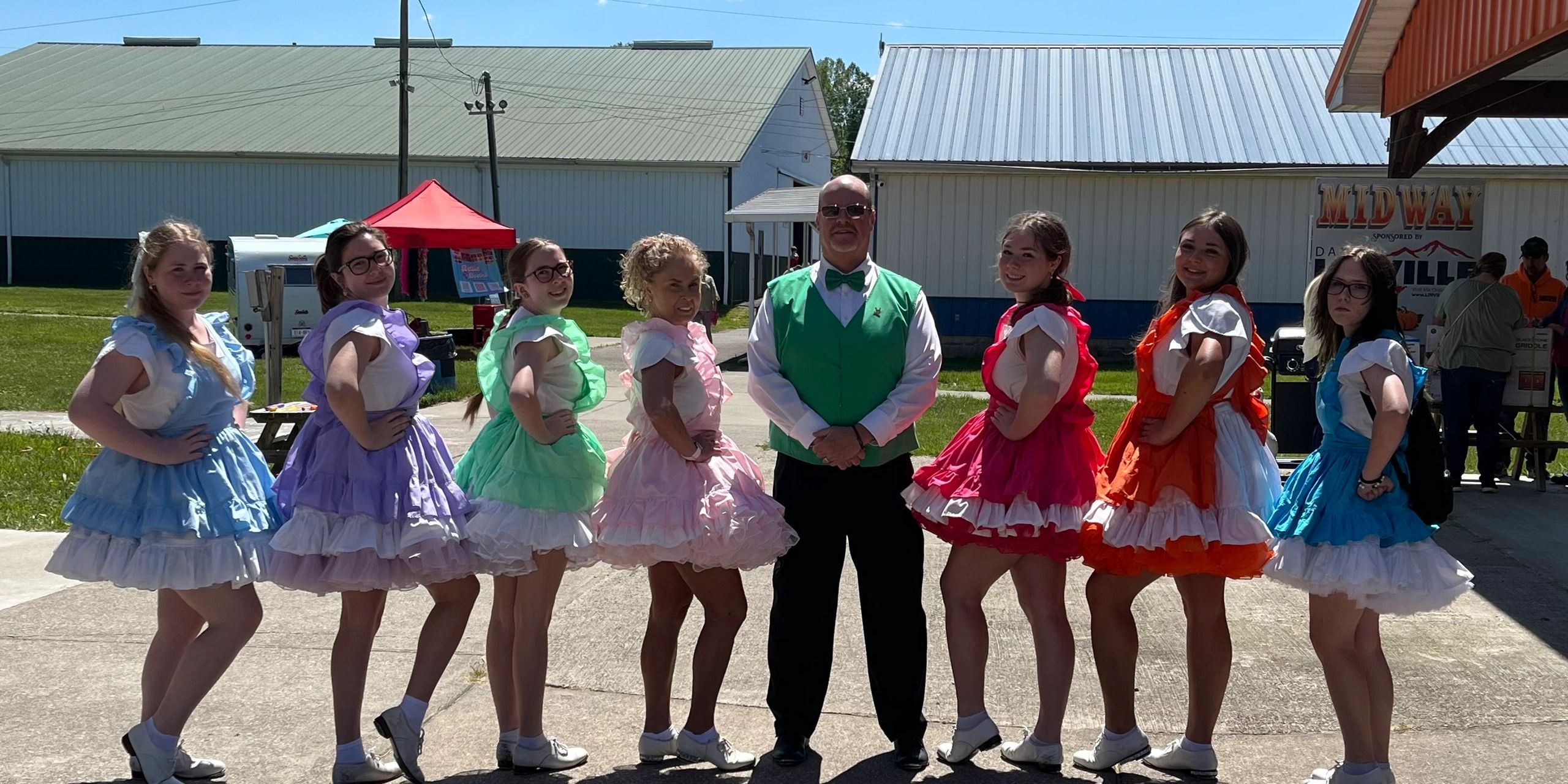 A group of dancers in colorful dresses posing outdoors with a man in a green vest.