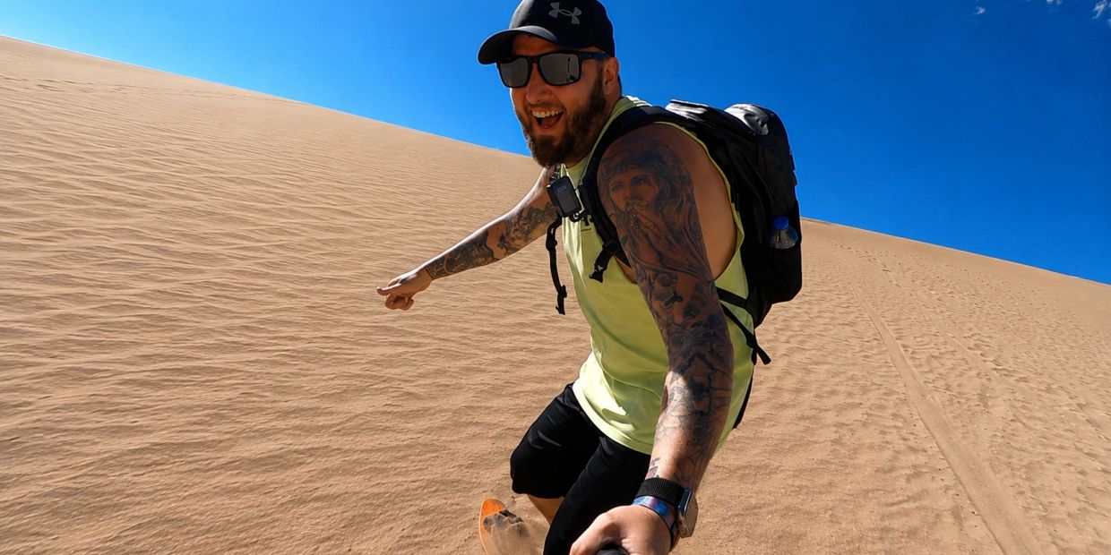 Man sandboarding on a sunny desert dune with tattoos and a backpack.
