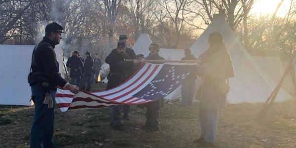 People in historical military uniforms holding a large American flag outdoors.