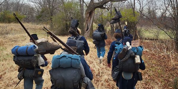 Group of Civil War reenactors marching through a field with backpacks and rifles.