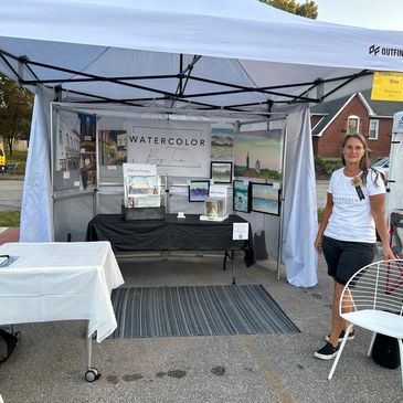 Woman standing by an art booth selling watercolor paintings.