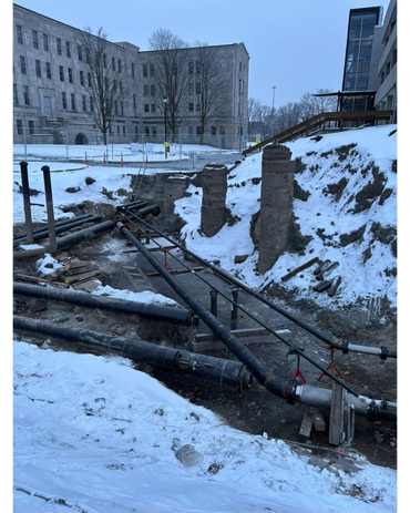 Construction site with exposed pipes and snow near large buildings.
