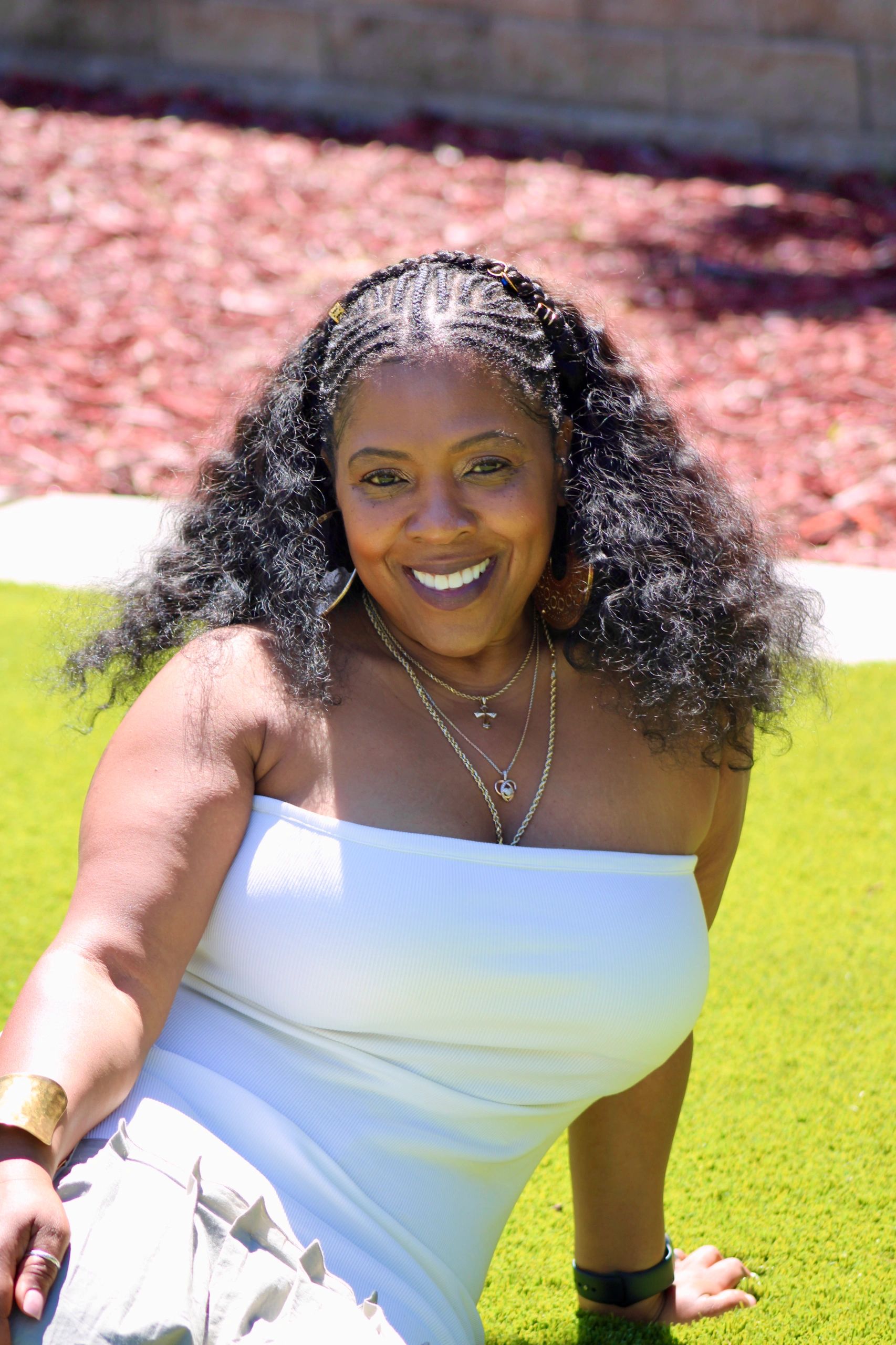 Smiling woman with curly hair in a white top sitting on green grass.