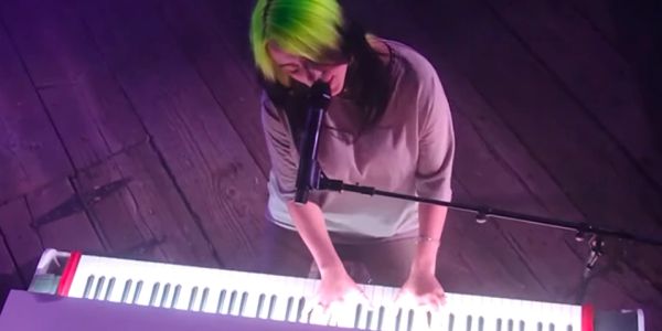 Singer with neon green hair plays keyboard under purple light.