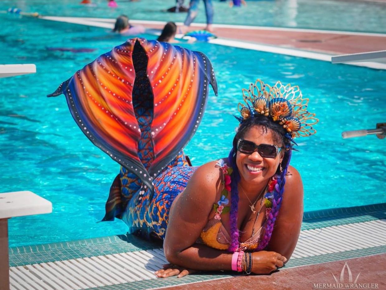 A woman in a colorful mermaid tail and crown poses by a pool, smiling brightly.