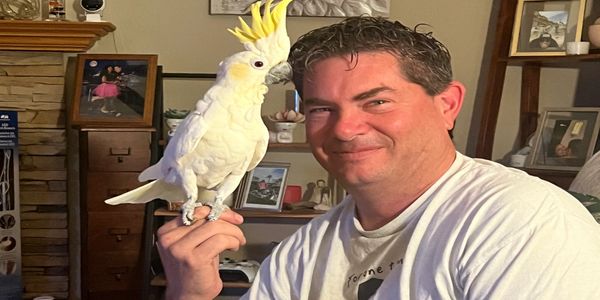 Man smiling while holding a white cockatoo on his finger indoors.