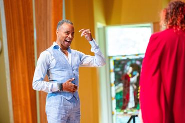 Man in light blue suit smiling and gesturing indoors.