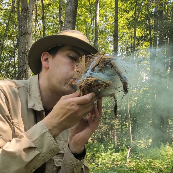 Man blowing on a smoky fire starter in the woods.