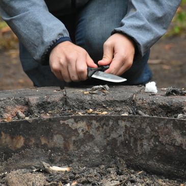 Hands holding a knife near a fire pit outdoors.