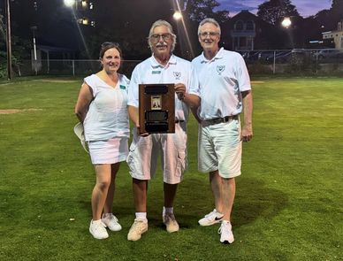 Three adults in white holding a trophy on a grass field at dusk.