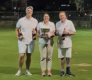 Three people in white holding trophies and champagne on a lawn at night.