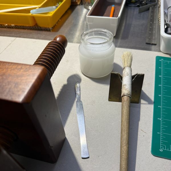 Bookbinding tools including a press, bone folder, brush, and jar of paste.