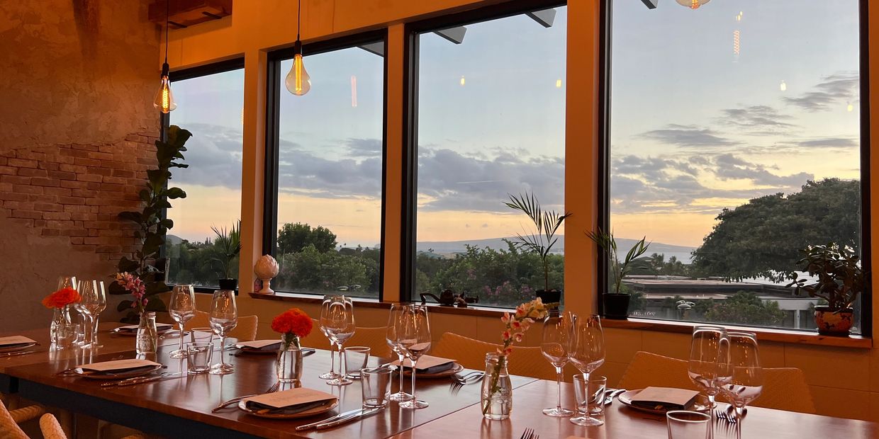 Elegant dining table set with flowers and glassware overlooking a sunset through large windows.