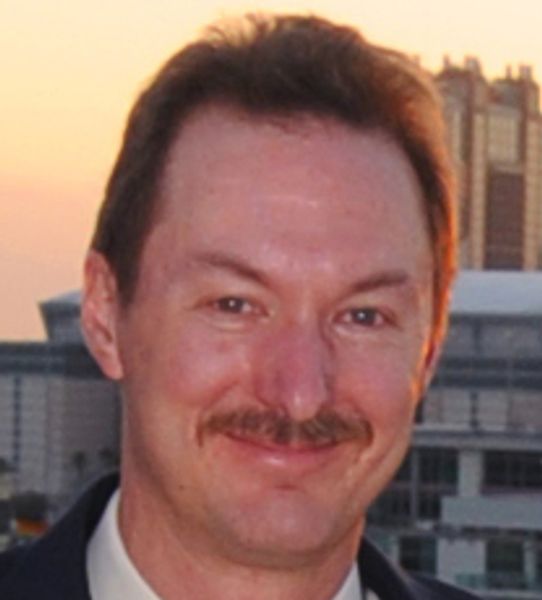 Man with a mustache smiling outdoors at sunset.