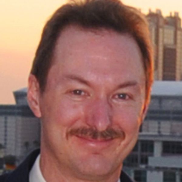 Man with a mustache smiling outdoors at sunset.