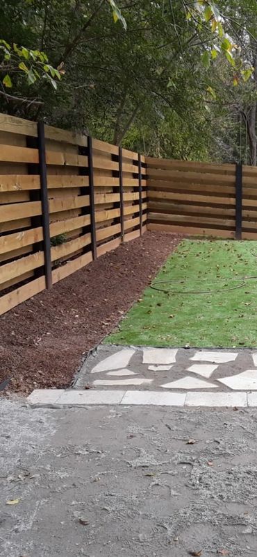 A backyard with a wooden fence, green grass, stone path, and mulch bed.