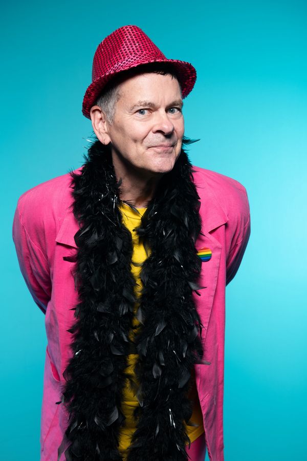 Man in pink suit and red hat with a black feather boa smiles against blue background.