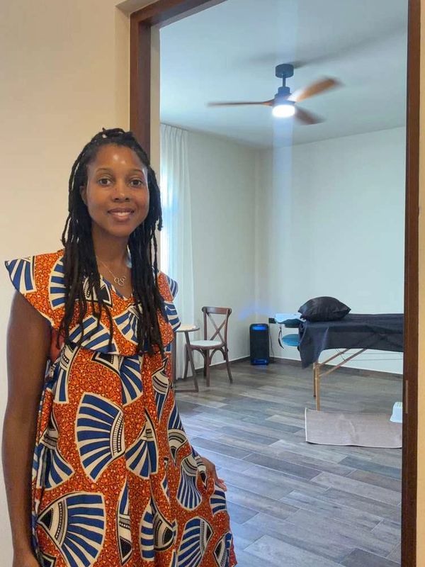 Mujer sonriendo en un vestido colorido, junto a una habitación con camilla y ventilador.