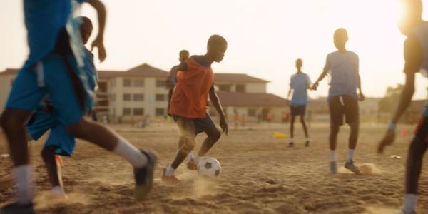 Children playing soccer on a dusty field during sunset.