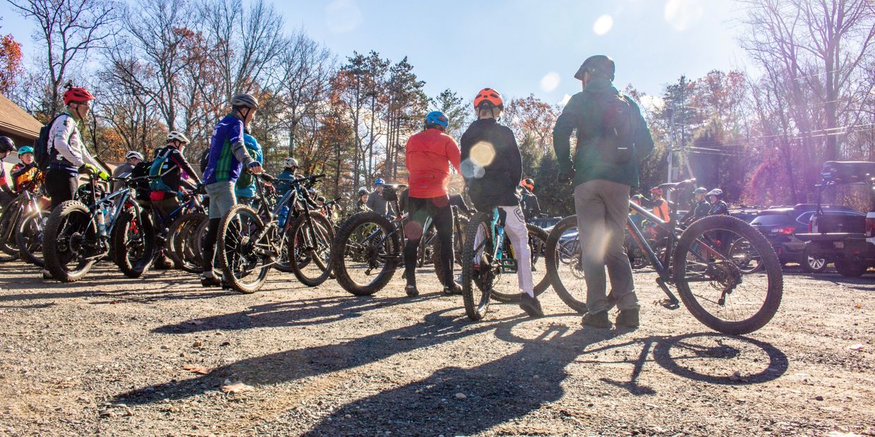A group of mountain bikers standing in a dirt lot with their bikes in the sun.