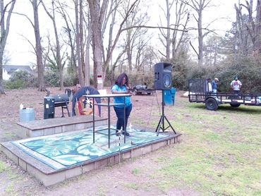 Woman speaking on a small outdoor stage in a wooded area.