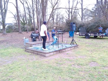 Two men on a small outdoor stage in a park area.