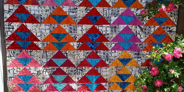 Colorful quilt with triangular patterns displayed outdoors beside blooming pink flowers.